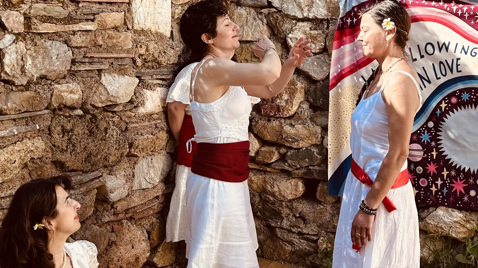 Beyaz elbiseli üç kadın Munay Ki töreninde, kozmik desenli pankart önünde