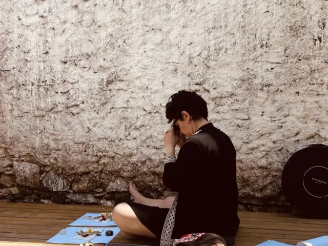 Özbilen Duygu taş duvar önünde şamanik objeler ve kristallerle hazırlık meditasyonunda